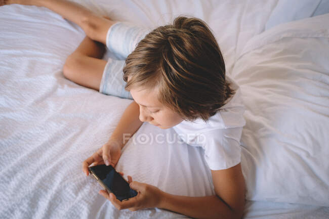 Boy in White Verifica o telemóvel de uma cama de quarto de hotel. — Fotografia de Stock