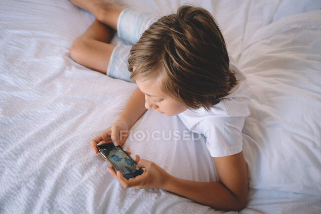 Junge in weißen Schecks spielt auf seinem Handy aus einem Hotelzimmerbett. — Stockfoto