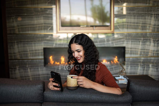 Mujer joven con taza de café y teléfono móvil en casa - foto de stock