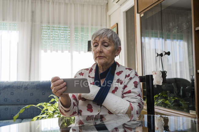 Femme âgée aux cheveux blancs caucasiens avec le bras cassé, assise à la maison — Photo de stock