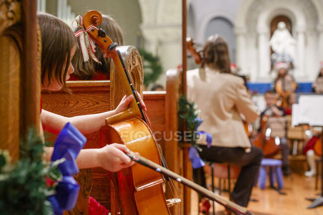 Una niña pequeña se sienta en el banco de la iglesia sosteniendo el violonchelo esperando para actuar - foto de stock