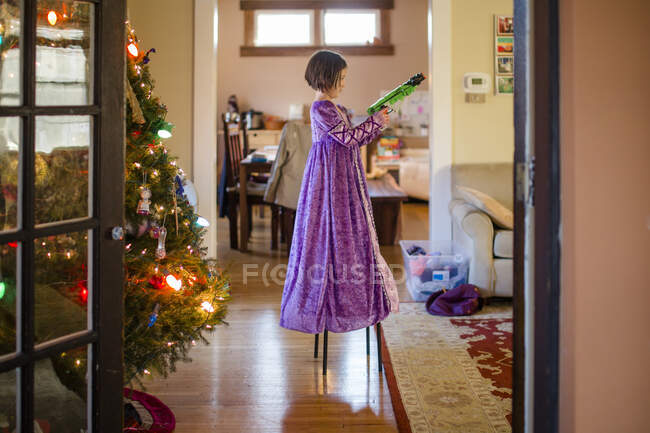 Un petit enfant dans une longue robe de princesse se tient sur une chaise avec pistolet jouet — Photo de stock