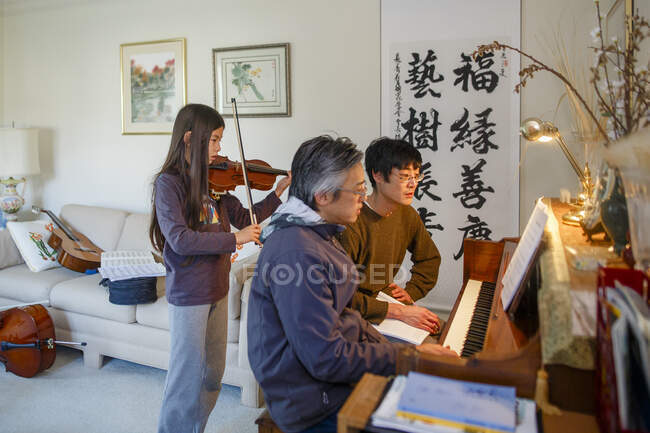 A father and uncle sit at piano while girl plays violin behind them — Stock Photo