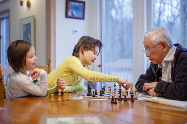 Un niño y su hermana se sientan en el tablero de ajedrez con el abuelo haciendo un movimiento - foto de stock