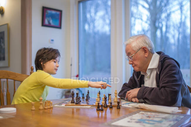 A boy makes a chess move while his grandfather watches on — Stock Photo