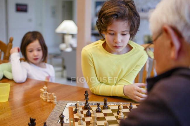Мальчик изучает шахматную доску, пока его сестра и дедушка смотрят — стоковое фото