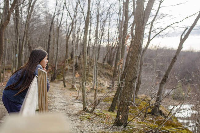 Una chica con el pelo largo se apoya en la valla mirando hacia fuera sobre un valle arbolado - foto de stock