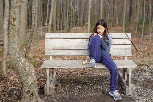 A tween girl sits alone on a bench in woods eating an orange in autumn — Stock Photo