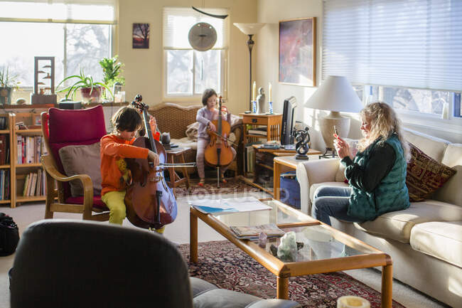 Two children perform cello at home and grandmother records with phone — Stock Photo