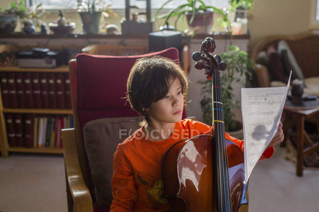 Un niño se sienta en una hermosa luz con un violonchelo leyendo partituras - foto de stock