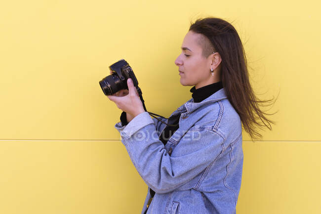 Young girl dressed in modern clothes uses her photo camera. Yellow wall background — Stock Photo