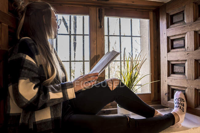 Жінка читає книгу, маючи чашку чаю біля вікна — стокове фото
