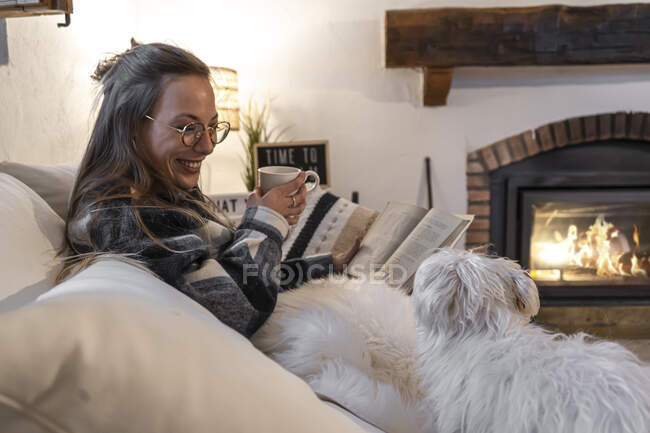 Glückliche Frau zu Hause, die auf einem Buch liegt und mit ihrem Hund Kaffee trinkt — Stockfoto