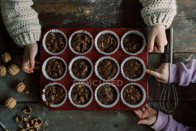 Manos de niño sosteniendo una bandeja de magdalenas de chocolate sin cocer a punto de hornear - foto de stock