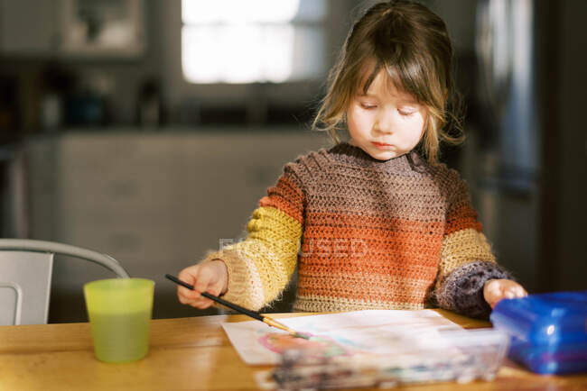 Маленькая дошкольница рисует акварелью за кухонным столом — стоковое фото