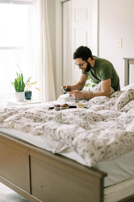 Man hacing breakfast on bed at  home and using smartphone — Stock Photo