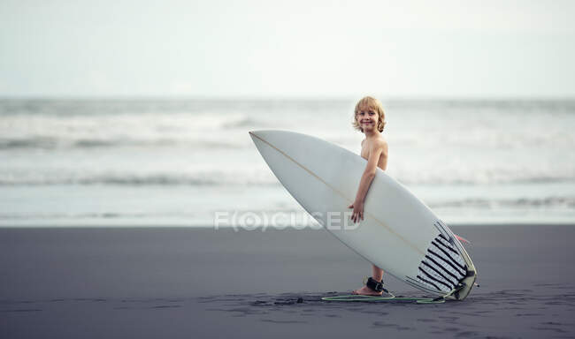 Niño pequeño cerca del océano con una tabla de surf sonriendo a la cámara - foto de stock