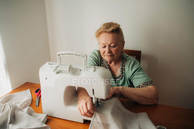 Femme âgée qui travaille sur une machine à coudre alors qu'elle est assise à la maison — Photo de stock