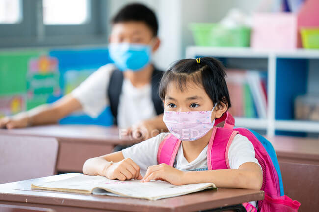 Niños de la escuela que usan máscara protectora para proteger contra Covid-19 con bolígrafos y cuadernos prueba de escritura en el aula, la educación, la escuela primaria, el aprendizaje y el concepto de personas. - foto de stock