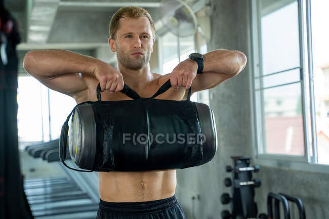 Starker Mann übt das Heben von Hanteltaschen, Muskulöser Fitnessmann macht Kniebeugen mit Fitnesstasche im Fitnessstudio. — Stockfoto