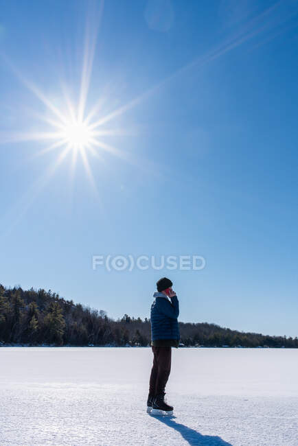 Мальчик-подросток на смартфоне и катается на коньках по замерзшему озеру в зимний день. — стоковое фото