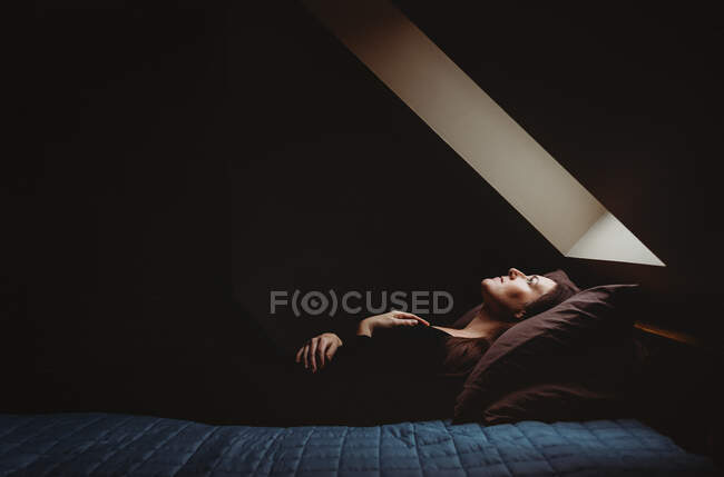 Mulher deitada em uma cama em um quarto escuro olhando para cima através de uma luz do céu. — Fotografia de Stock
