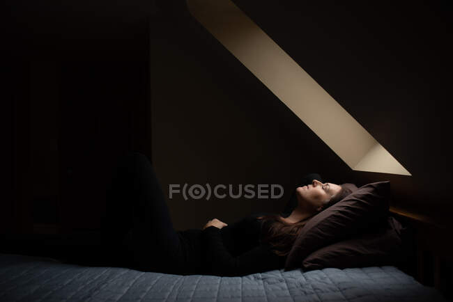 Frau liegt mit geschlossenen Augen im dunklen Raum unter einem Himmelslicht auf dem Bett. — Stockfoto