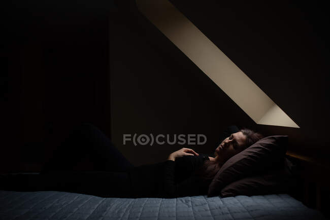 Donna che posa su letto con occhi chiusi in camera scura sotto una luce di cielo. — Foto stock