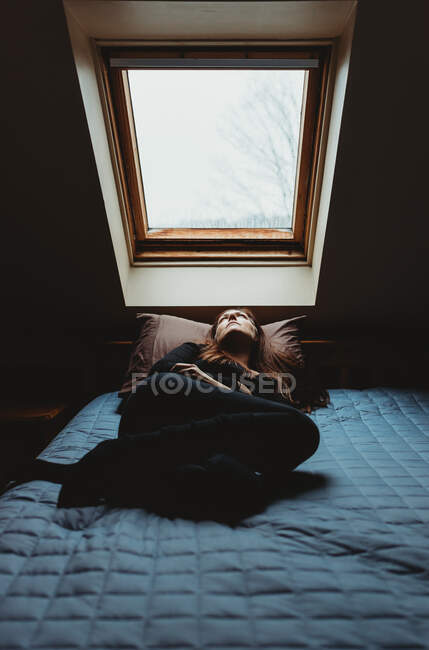 Frau liegt in einem dunklen Raum auf dem Bett und blickt durch ein Himmelslicht nach oben. — Stockfoto