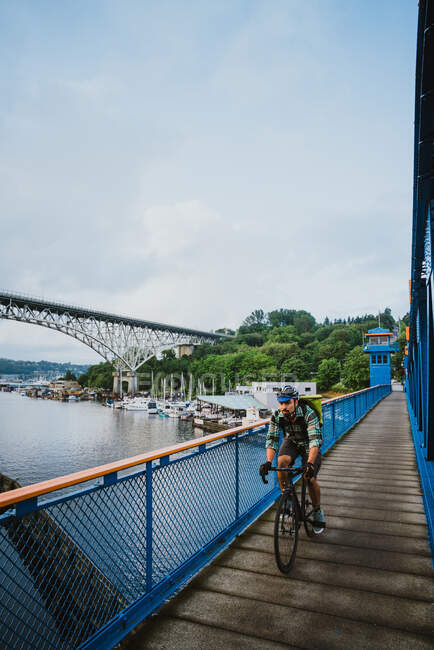 A young man bikes across a bridge with water and boats in the distance — Stock Photo