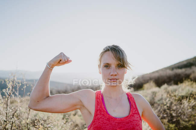 Mulher loira atlética flexiona músculos com sol e montanhas atrás — Fotografia de Stock