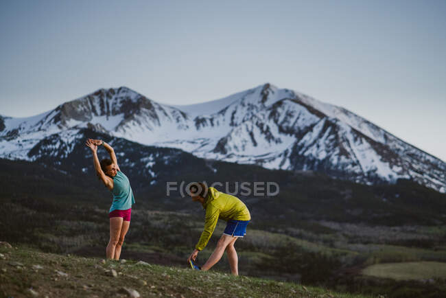 Two friends stretch before trail running with large mountains in view — Stock Photo