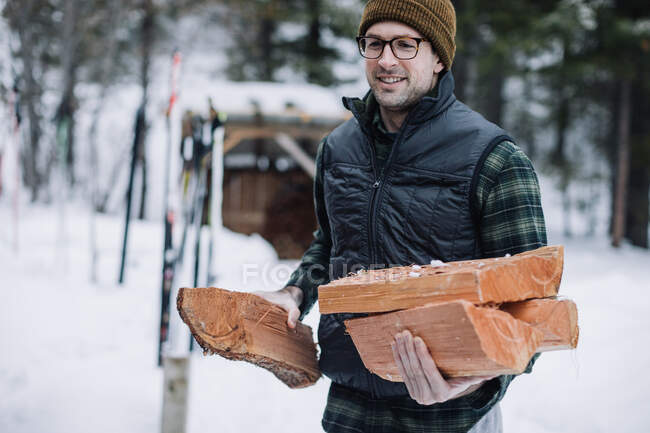 Молодой человек в очках и клетчатой клетке несет дрова обратно в лыжную хижину — стоковое фото
