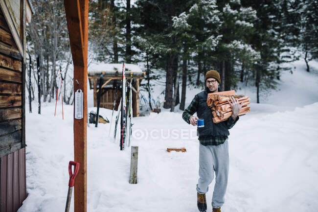 A man wearing flannel carries firewood and a coffee back to cabin — Stock Photo