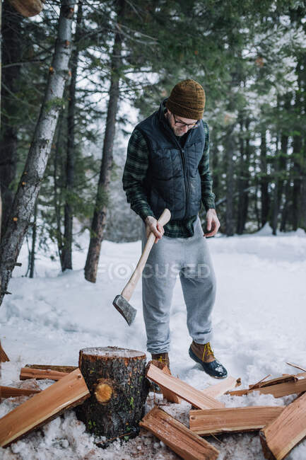 A man wearing beanie and flannel chops firewood in the snow — Stock Photo