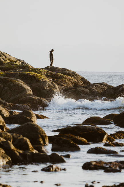 Un hombre ve la ola estrellarse contra la costa rocosa durante la marea baja - foto de stock