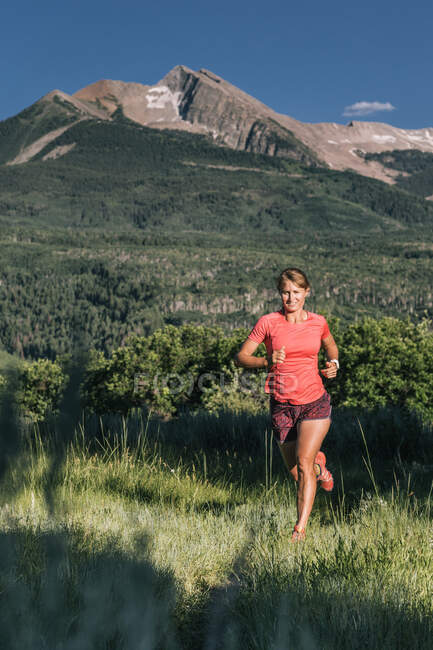 Trilha de mulher atlética atravessa campo gramado com vista épica da montanha — Fotografia de Stock
