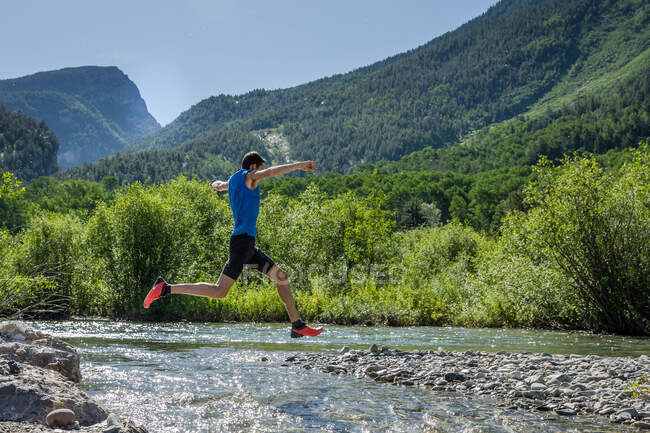 Masculino trilha corredor salta sobre rochoso rio no ensolarado dia no o montanhas — Fotografia de Stock