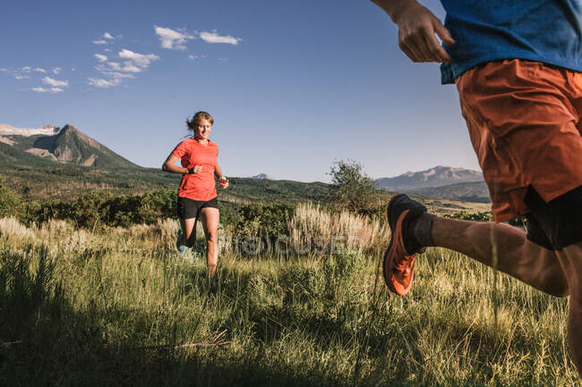 Dos corredores de senderos corren a través de un campo de hierba alta con vista a la montaña - foto de stock