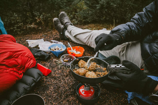 A man cooks dinner on the ground while backpacking wearing gloves — Stock Photo