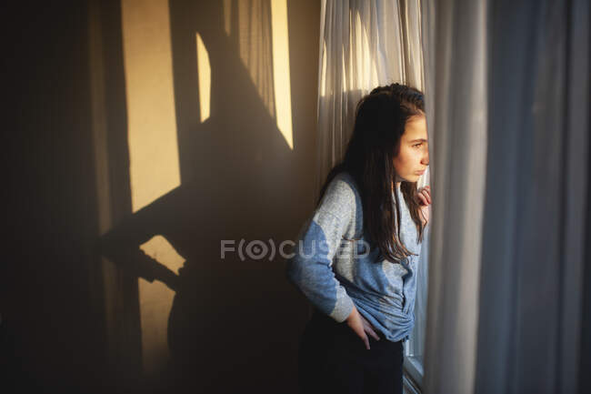 Adolescente regarde par une fenêtre ensoleillée avec expression concernée — Photo de stock