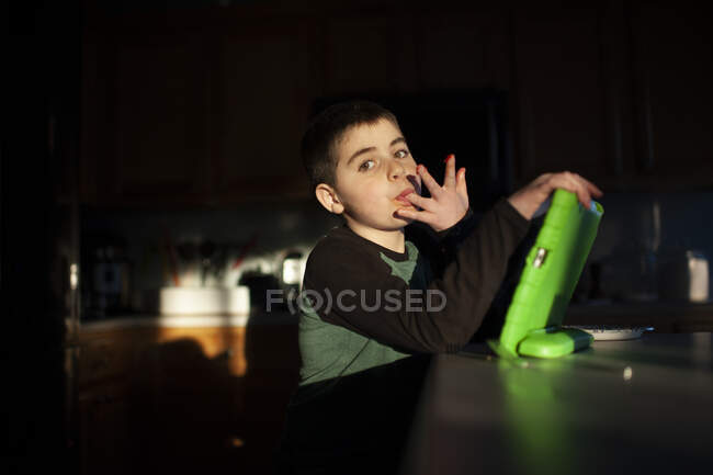 Menino 9-10 anos de idade lambe os dedos enquanto segurando tablet em luz bonita — Fotografia de Stock