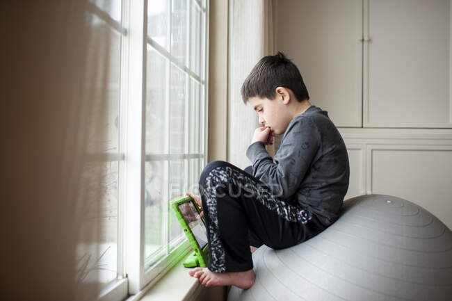 Garçon 9-10 ans est assis sur la balle d'exercice en jouant avec sa tablette — Photo de stock