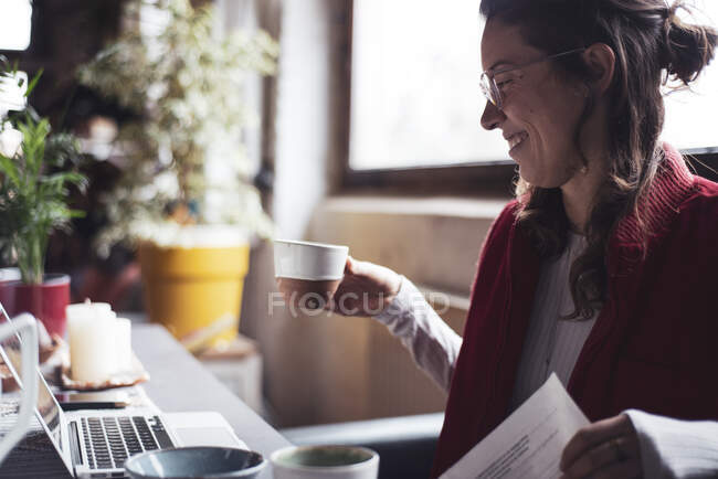 Femme sourit avec une tasse de thé tout en travaillant sur ordinateur portable au bureau à la maison — Photo de stock