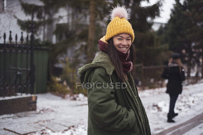 Счастливая азиатка с желтой пушистой шапочкой улыбается на снежной улице — стоковое фото