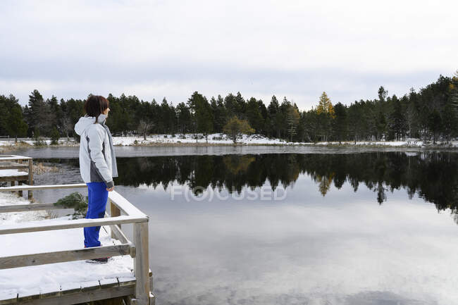 Femme debout sur un pont en bois enneigé regardant vers un lac gelé — Photo de stock