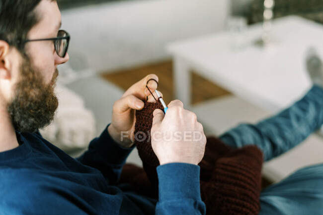 Millennial hombre tejiendo un suéter de niño en un sofá en la sala de estar - foto de stock