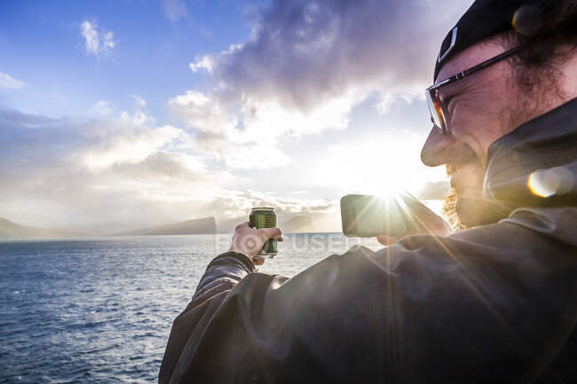 Pasajero del barco sosteniendo una lata de bebida mientras toma una fotografía en su teléfono durante la puesta del sol después de salir del puerto de Torshavn en las Islas Feroe - foto de stock