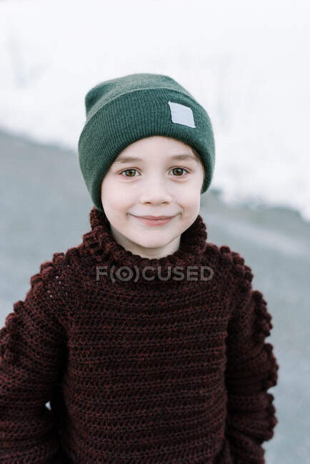 Petit garçon souriant avec chapeau dans un pull fait maison debout à l'extérieur — Photo de stock