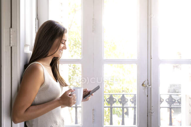 Young woman using mobile phone while sitting on windowsill at home — Stock Photo